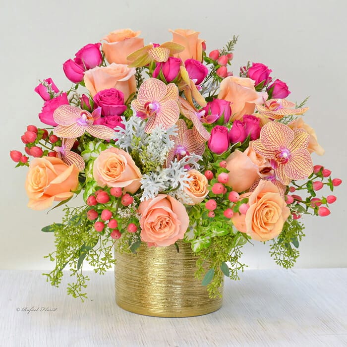 Introducing "Carnival - B91": a luxurious floral arrangement featuring peach and pink roses, complemented by orchids, vibrant green foliage, and red berries, all beautifully arranged in a gold textured vase. Perfect for adding a touch of elegance to any space or as a thoughtful gift for someone special.